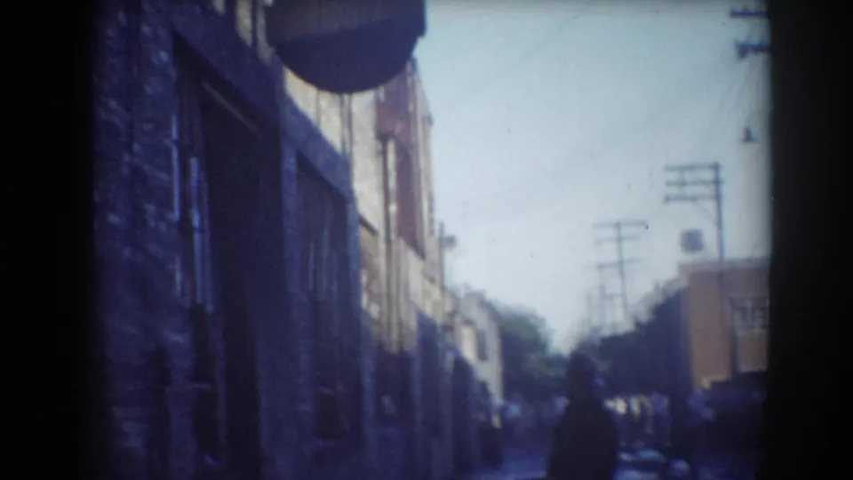 Blue-cast 8mm frame looking along a York, Nebraska street, with brick building wall, overhead lines and people near the far end.