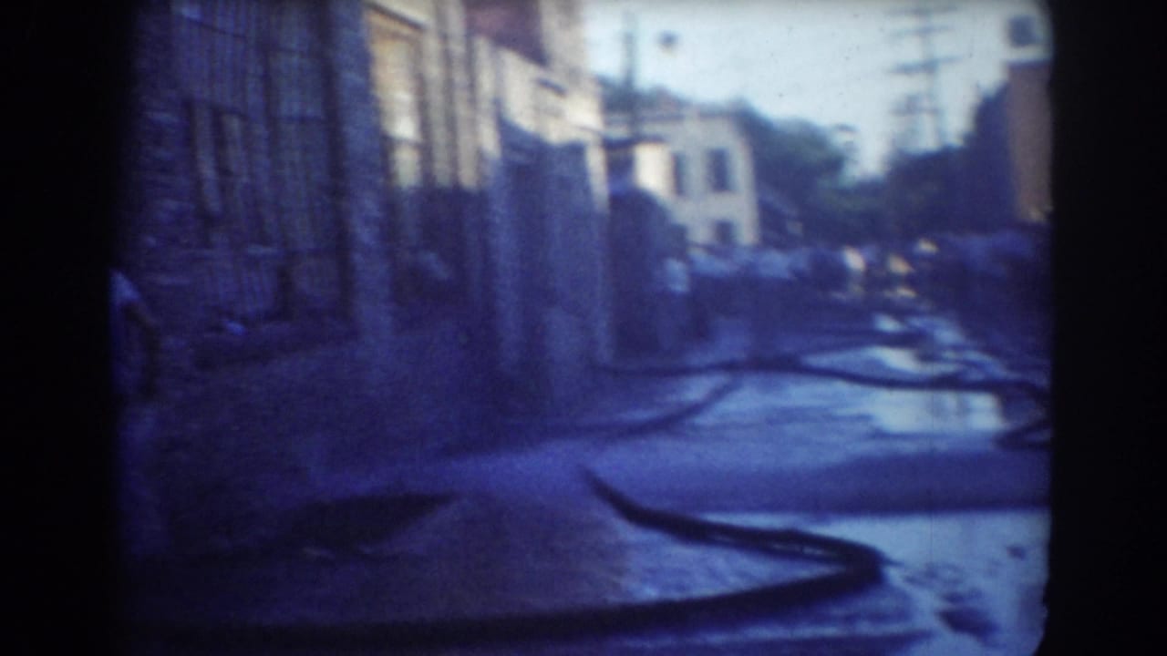 Wet urban street with hose running across pavement toward a distant crowd.
