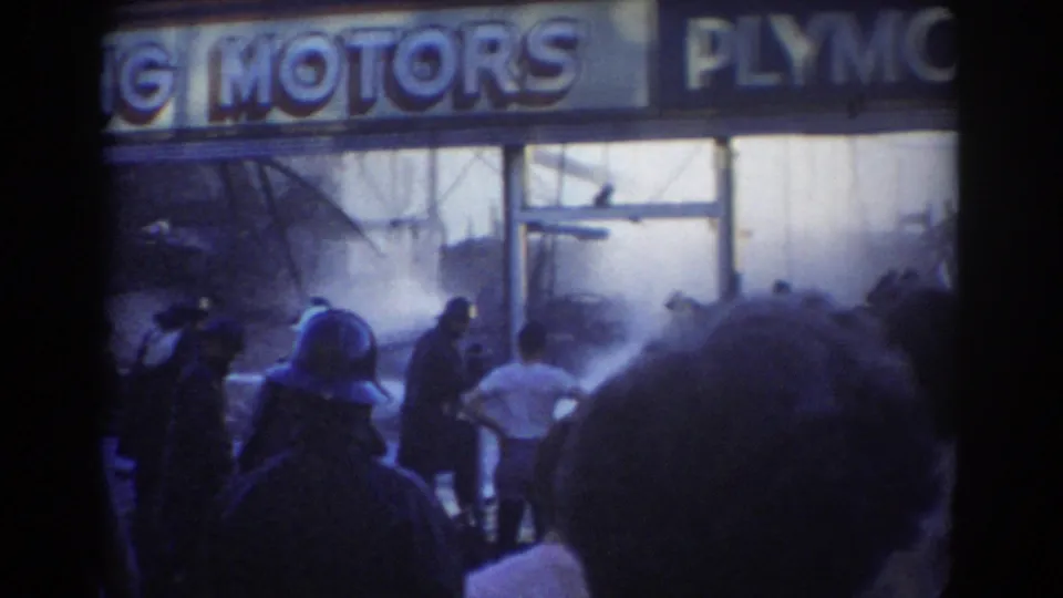 1958 York, Nebraska 8mm frame showing bystanders facing a smoky Plymouth Motors dealership as responders work near the front windows.