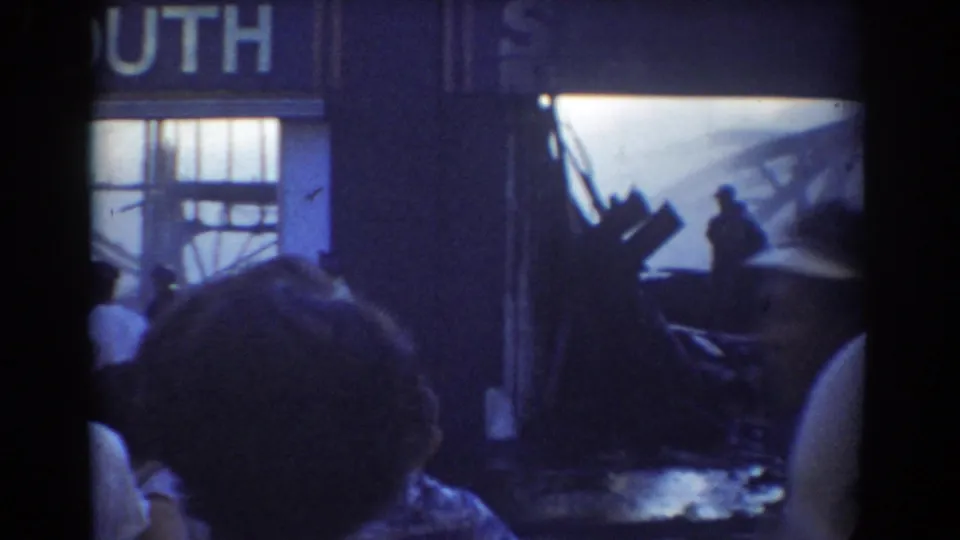 Late frame from the York, Nebraska Plymouth Motors fire showing smoke, a silhouetted responder or bystander, debris, and crowd heads at the edge.