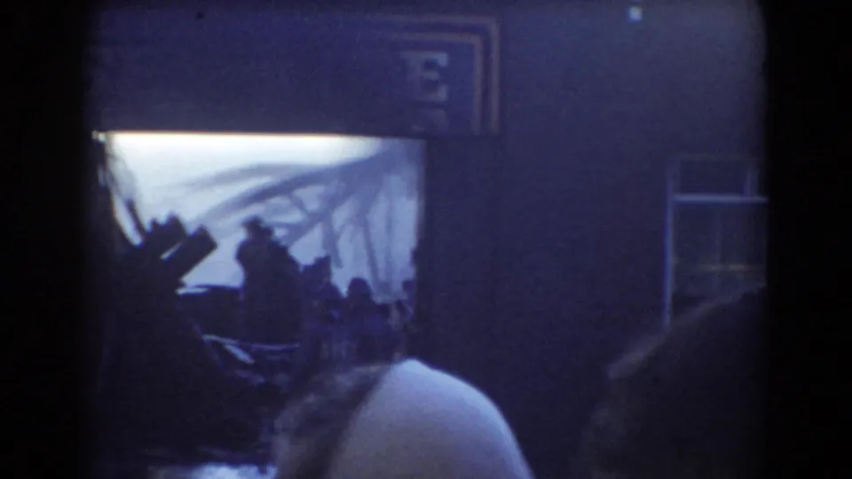 Crowd members in a 1958 York, Nebraska 8mm frame watch activity near a storefront with partial signage and bright haze beyond.