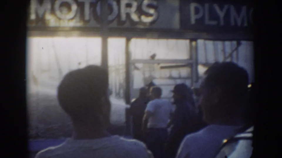 1958 York, Nebraska 8mm frame showing spectators from behind facing a storefront with MOTORS and partial PLYMOUTH lettering.