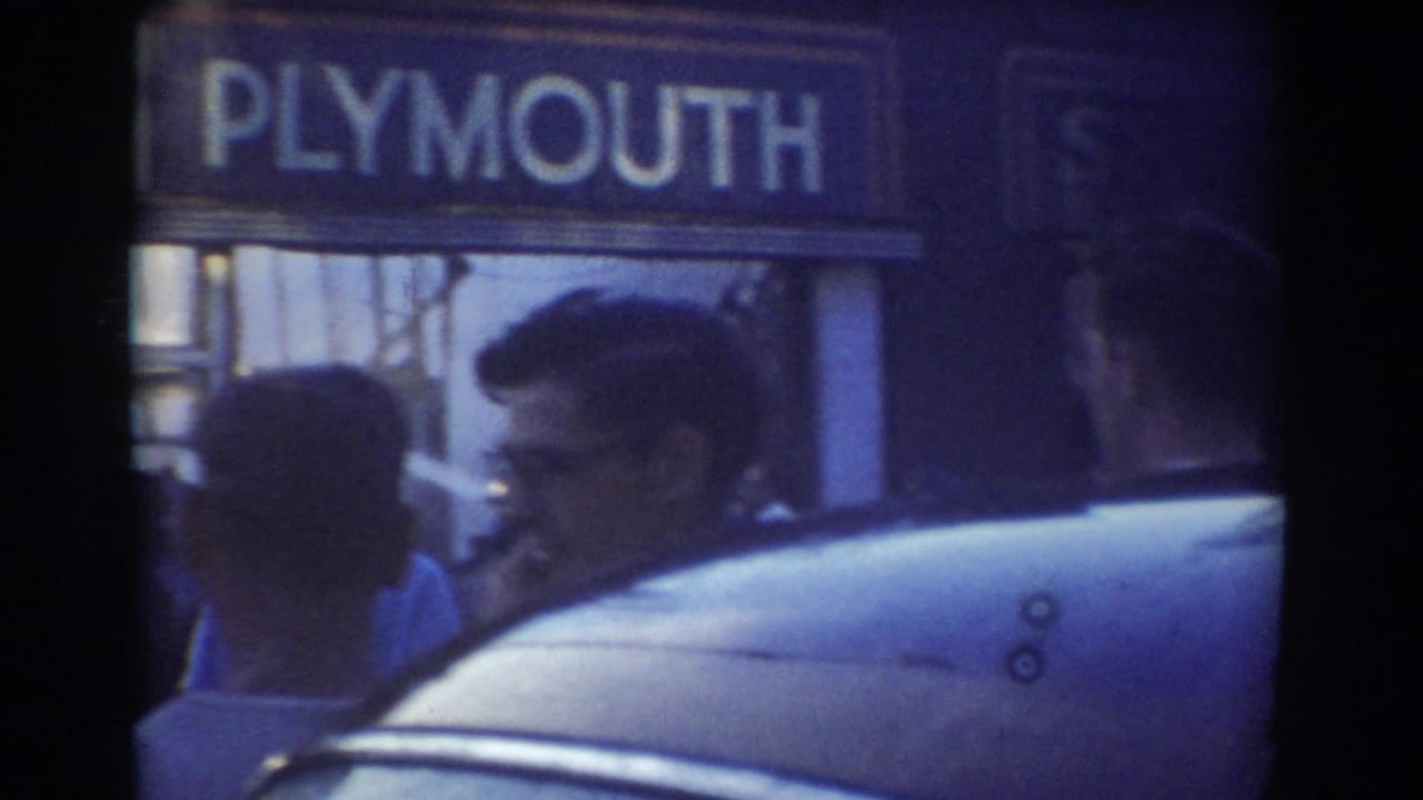 1958 York, Nebraska frame with a Plymouth sign above the sidewalk, several adults in profile, and the roofline of a parked car in the foreground.