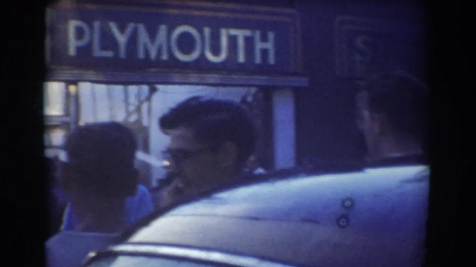 1958 York, Nebraska frame with a Plymouth sign above the sidewalk, several adults in profile, and the roofline of a parked car in the foreground.