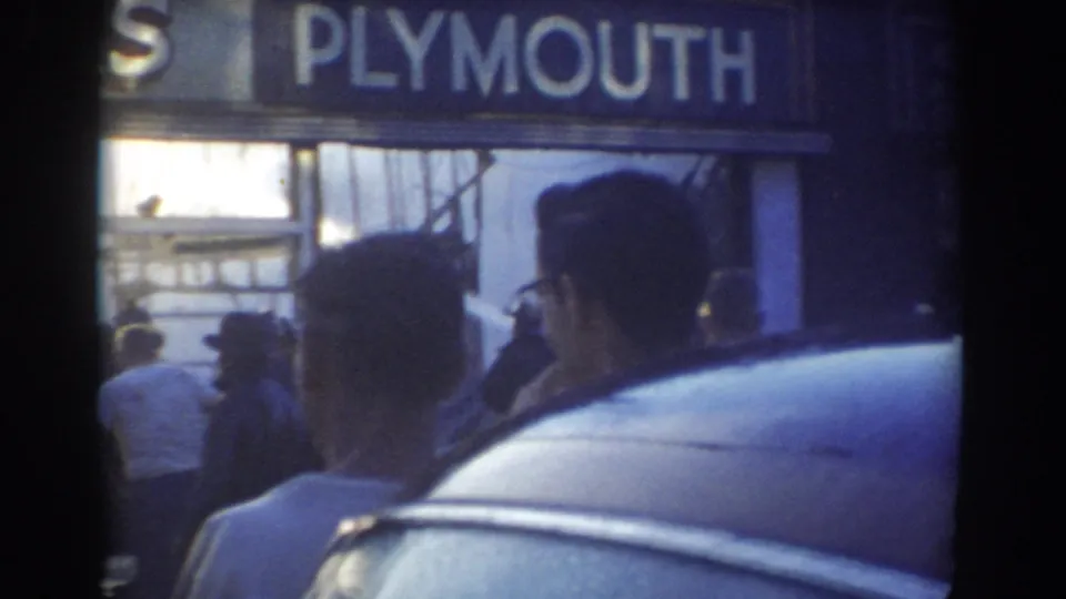 1958 York, Nebraska home-movie still showing men near a parked car and a large Plymouth sign above the dealership frontage.