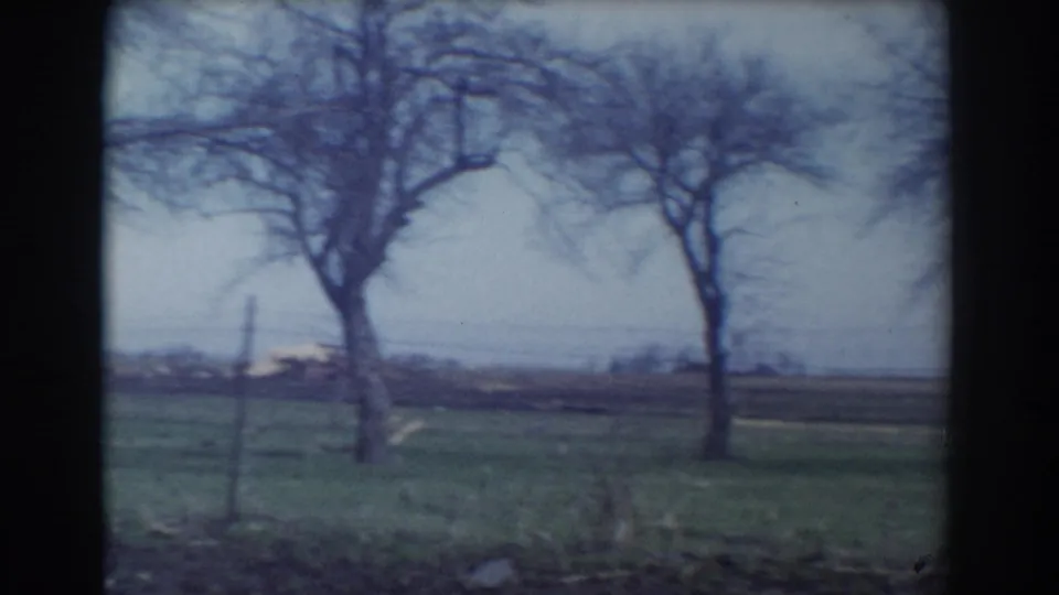 York, Nebraska home-movie frame showing bare trees, fence posts, grass, and distant open fields under a muted sky.
