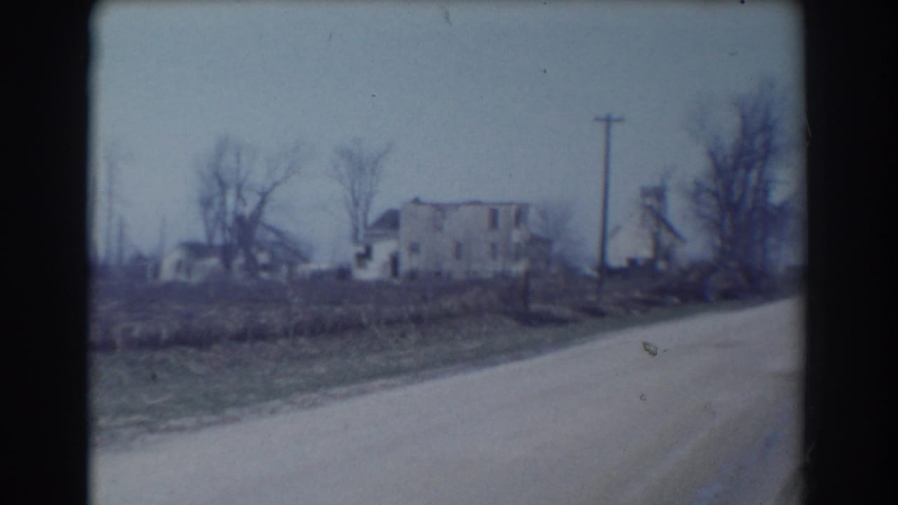 1958 York, Nebraska 8mm frame showing a pale rural road curving past a weathered two-story house, bare trees, grassy roadside, and utility poles.