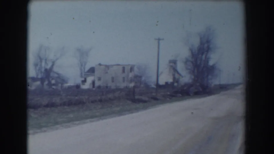 1958 York, Nebraska roadside view with an unpaved or light-colored rural road, a distant house, bare trees, utility poles, and open ground under a pale sky.