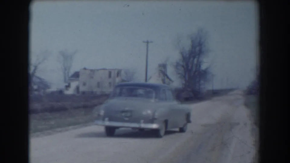 1958 York, Nebraska frame showing a mid-century sedan moving along the roadside, with a weathered house, bare trees, utility pole, and open rural landscape behind it.