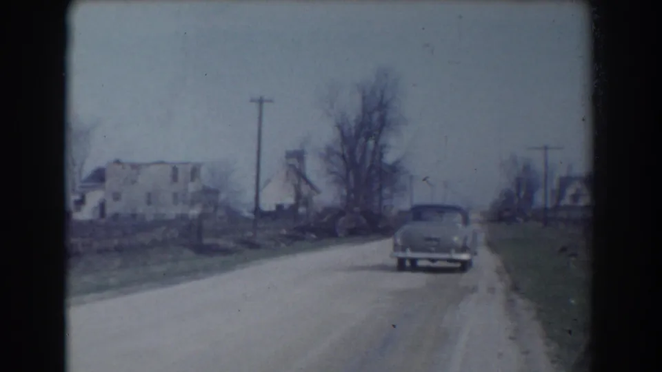 1958 York, Nebraska 8mm frame of a mid-century sedan receding down a rural road, with bare trees, utility poles, grassy shoulders, and a weathered roadside house visible.