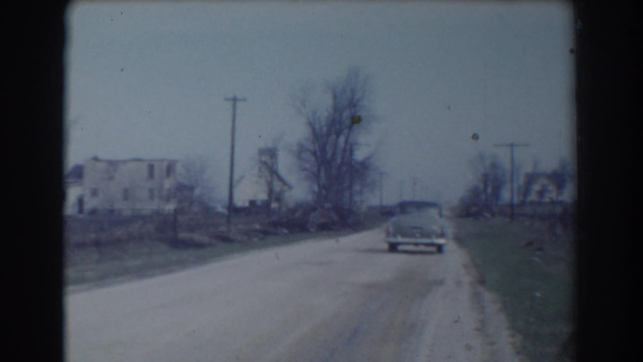 Vintage car driving away on a pale rural dirt road with houses, bare trees, and utility poles.