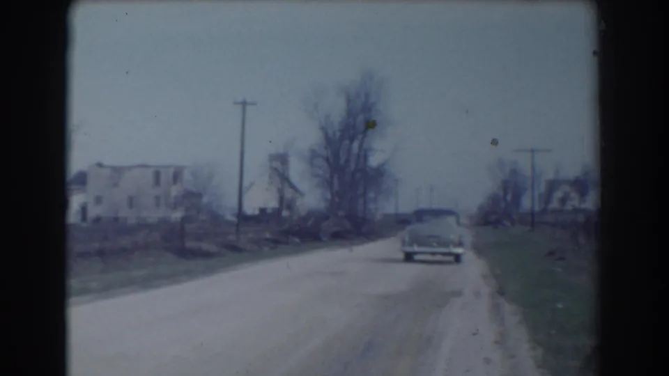 In York, Nebraska in 1958, a vintage car moves away from the camera along a dirt road bordered by houses, bare trees, grass, and utility poles.
