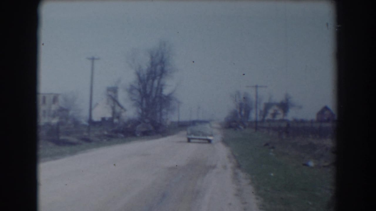 The 1958 York, Nebraska home-movie frame shows the car continuing away on the unpaved road past utility poles, bare trees, and scattered houses.