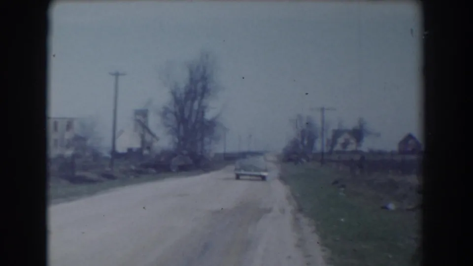 The 1958 York, Nebraska home-movie frame shows the car continuing away on the unpaved road past utility poles, bare trees, and scattered houses.