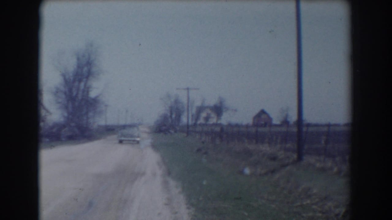 The car appears small in the distance on a rural York, Nebraska road, with roadside grass, fencing, utility poles, bare trees, and houses visible under a gray sky.