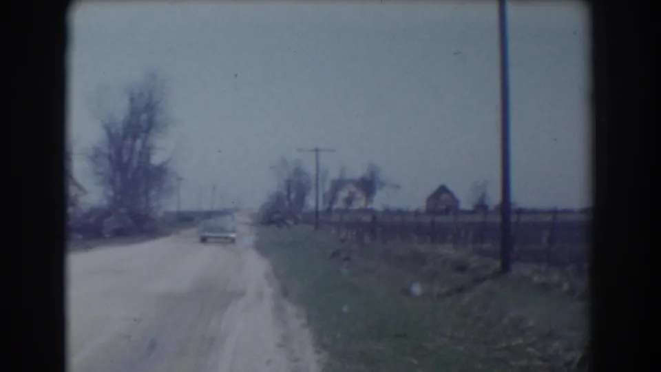 The car appears small in the distance on a rural York, Nebraska road, with roadside grass, fencing, utility poles, bare trees, and houses visible under a gray sky.