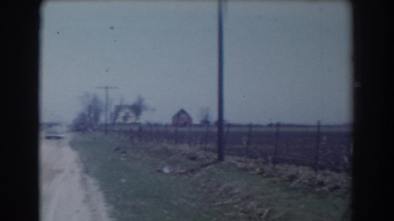 Near the end of the York, Nebraska clip, the car is almost out of view while the frame holds the road shoulder, fence line, utility pole, fields, and distant houses.