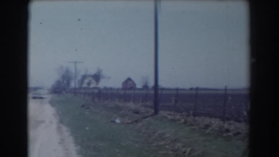 Near the end of the York, Nebraska clip, the car is almost out of view while the frame holds the road shoulder, fence line, utility pole, fields, and distant houses.