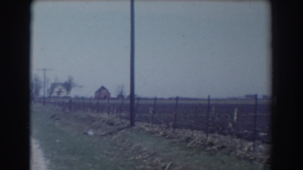 Dirt road beside a wire fence with distant rural buildings under an overcast sky.