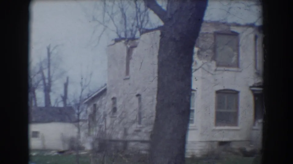 1958 York, Nebraska frame showing a weathered house wall, shuttered or dark windows, bare branches, and a large foreground tree.