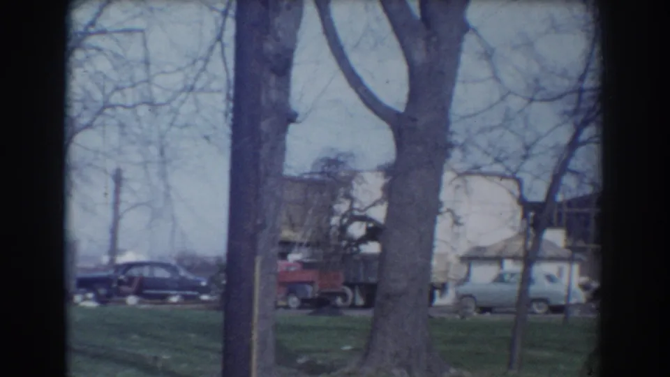 1958 York, Nebraska home-movie view across grass and tree trunks toward parked cars and residential buildings.