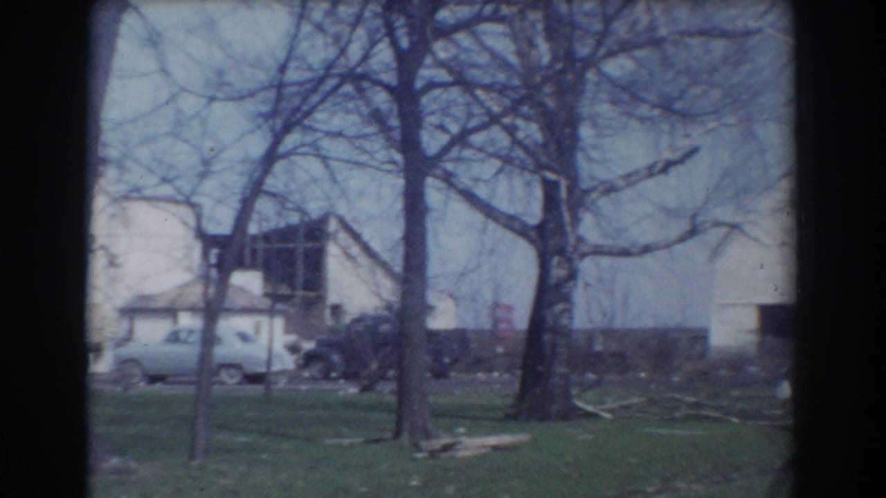 1958 York, Nebraska 8mm frame showing a rural farmyard with white outbuildings, leafless trees, parked cars, and debris on the grass.