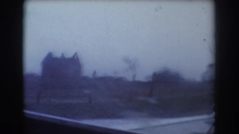 Soft 8mm car POV from York, Nebraska, showing a rural road edge, open ground, and a dark distant house silhouette.