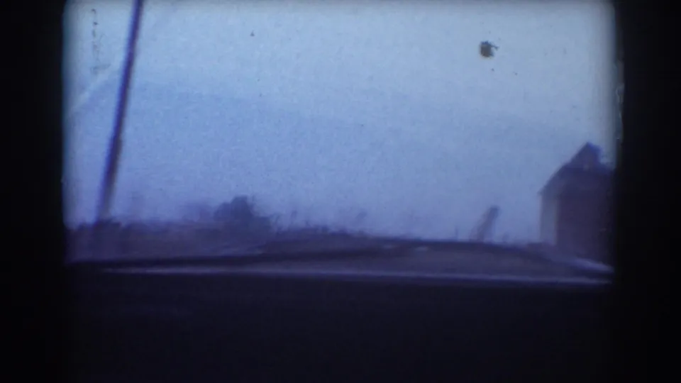 1958 York, Nebraska frame showing the roadside, a partial house at the right edge, bare vegetation, and the dark lower edge of the car window.