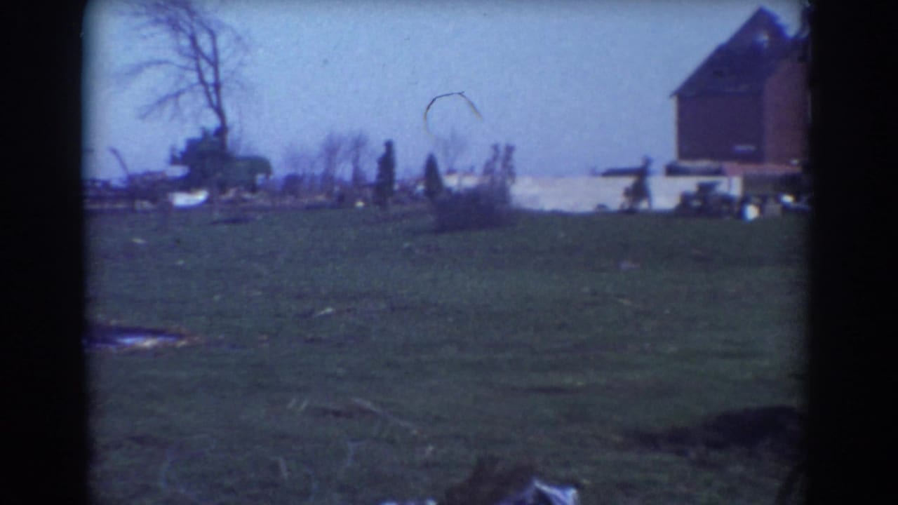 Grassy field with a damaged rural building, bare tree, debris, and distant people.