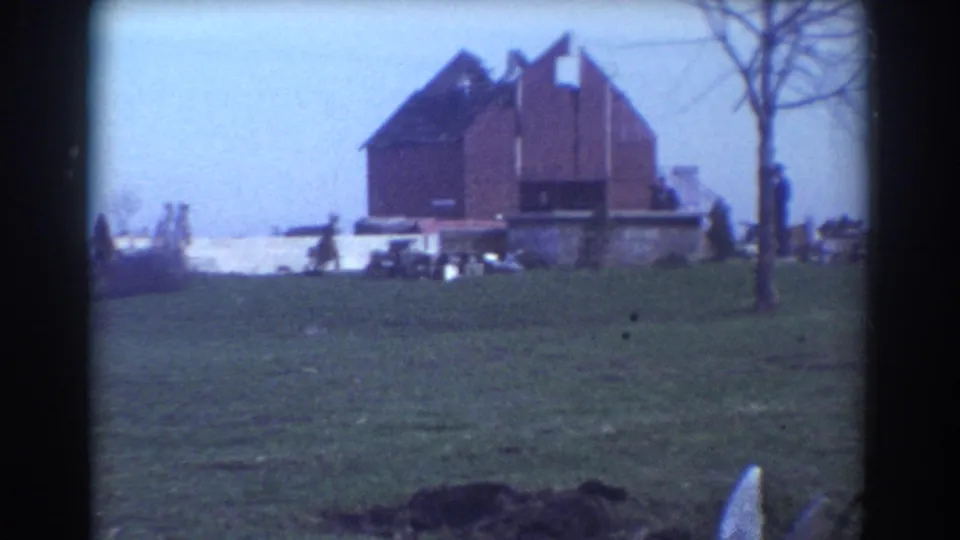 York, Nebraska 1958 home-movie frame of a damaged red barn-like building beside open grass and scattered storm debris.