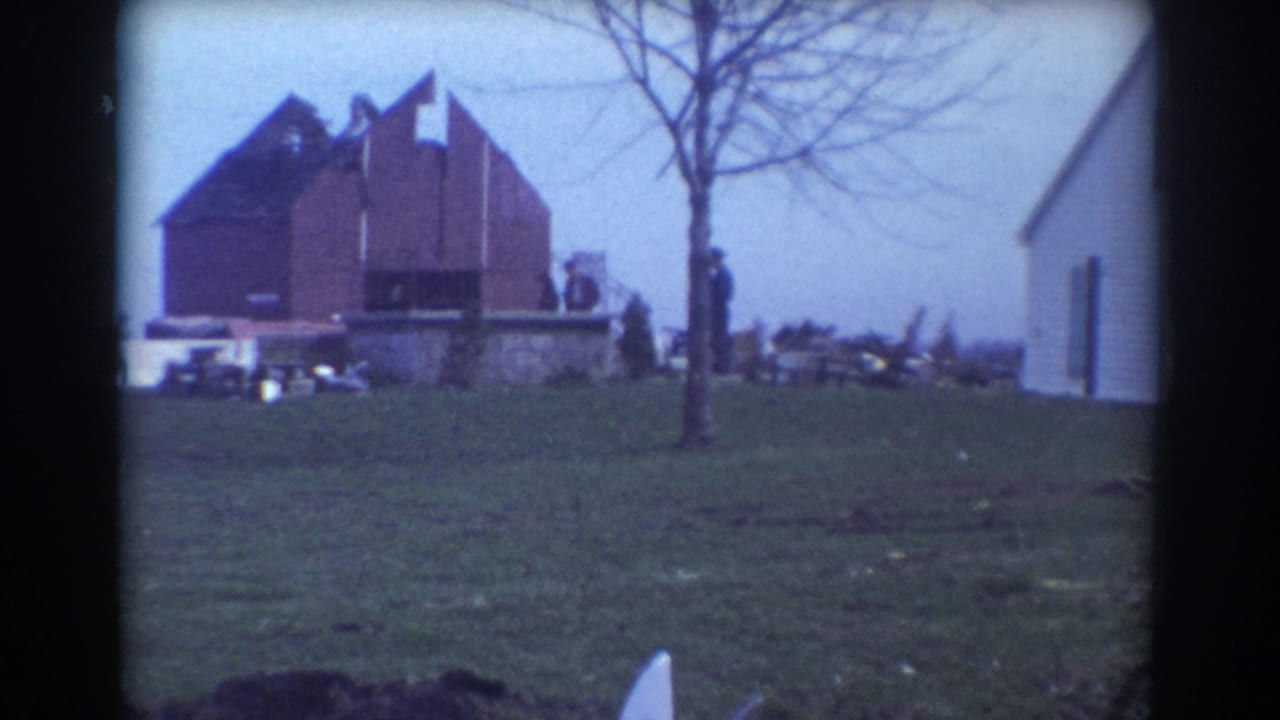 1958 York, Nebraska 8mm frame showing a red brick structure, part of a white house, bare tree, grassy yard, and small distant figures.