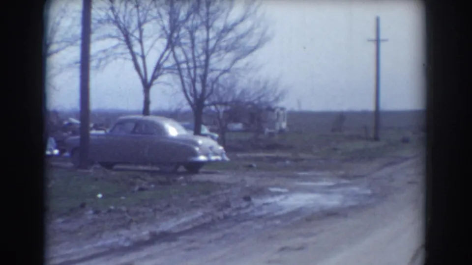1958 York, Nebraska frame showing a pale rounded car parked beside a muddy road, with bare trees, a utility pole, grass, and open land.