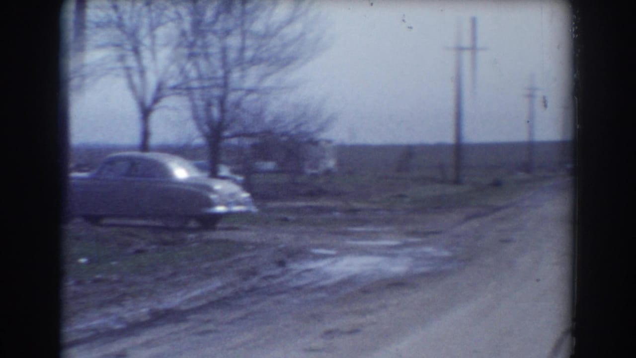 1958 York, Nebraska home-movie frame showing a muddy roadside, older car, utility poles, bare trees, and open rural fields after storm damage.