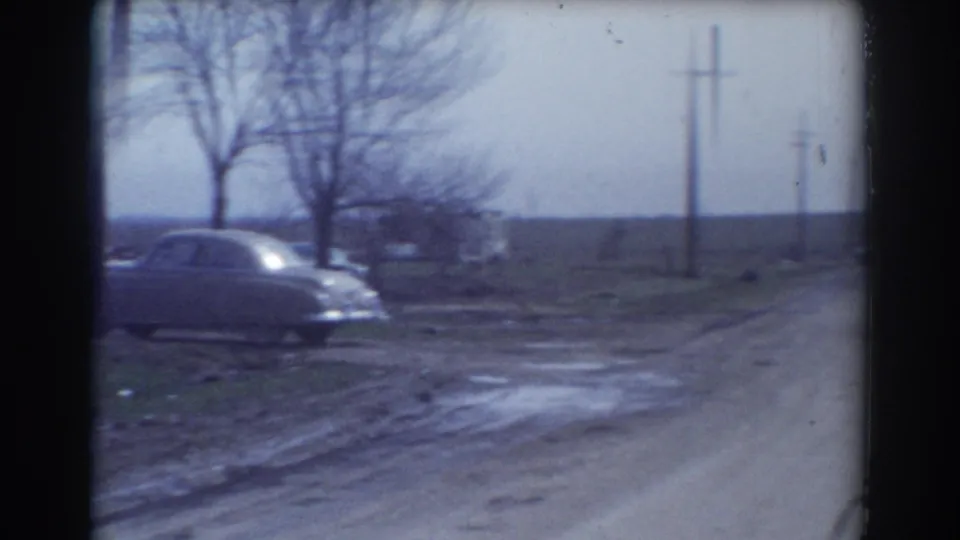 1958 York, Nebraska home-movie frame showing a muddy roadside, older car, utility poles, bare trees, and open rural fields after storm damage.