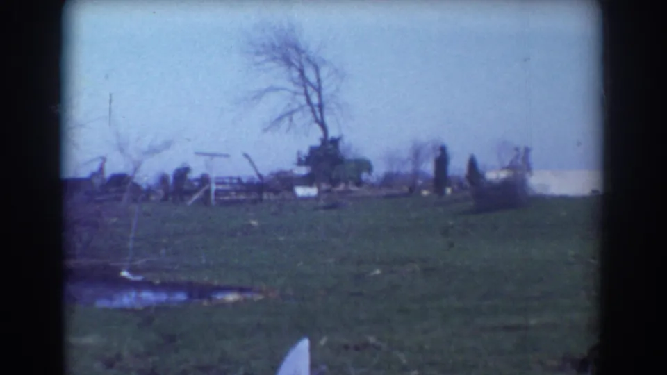 1958 York, Nebraska home-movie view of an open field with debris, bare trees, and distant damaged rural structures after a tornado.