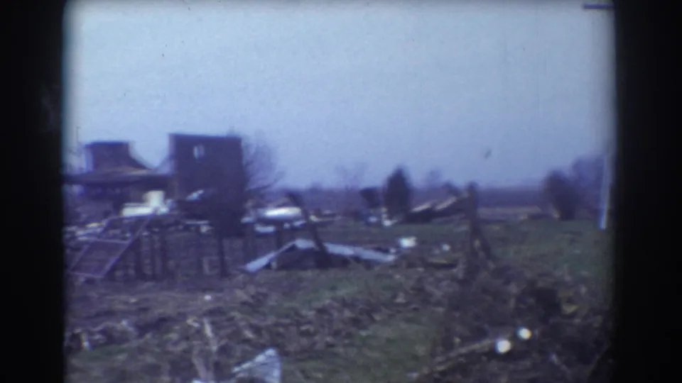 1958 York, Nebraska frame showing a damaged rural building area, scattered wreckage, a vehicle, bare trees, and open fields after tornado damage.