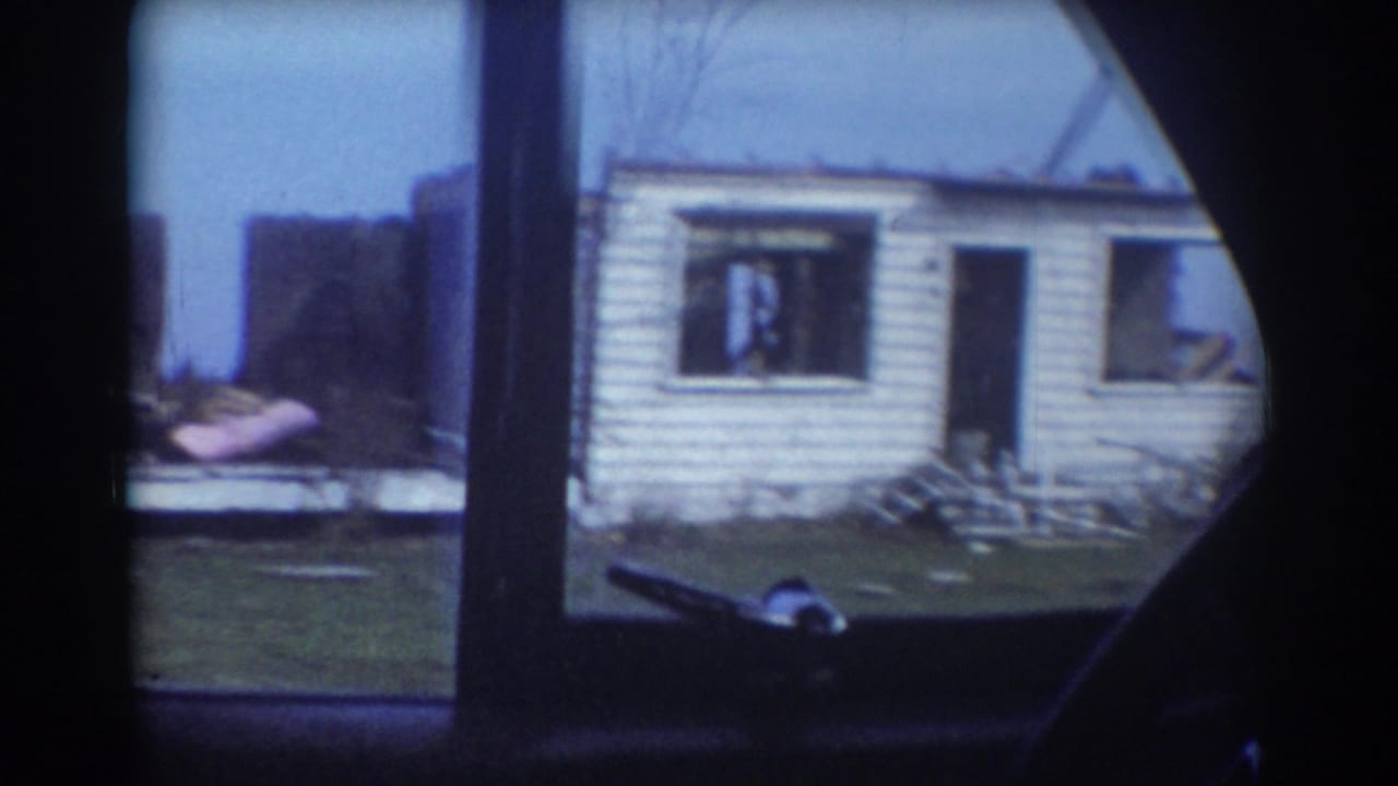 Damaged white-sided house seen through a vehicle window or frame edge.