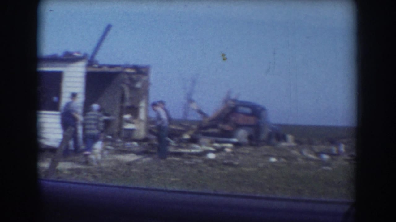 Several adults stand beside a damaged structure, debris, and a wrecked vehicle or equipment.