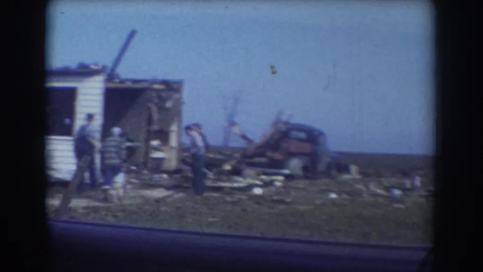 York, Nebraska storm aftermath frame with adults surveying debris beside a damaged building and wrecked vehicle or equipment.