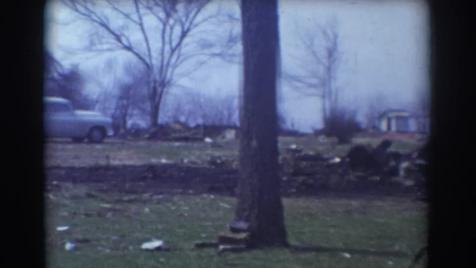 1958 York, Nebraska frame with a light mid-century car, bare trees, a foreground trunk, and debris scattered along the roadside.