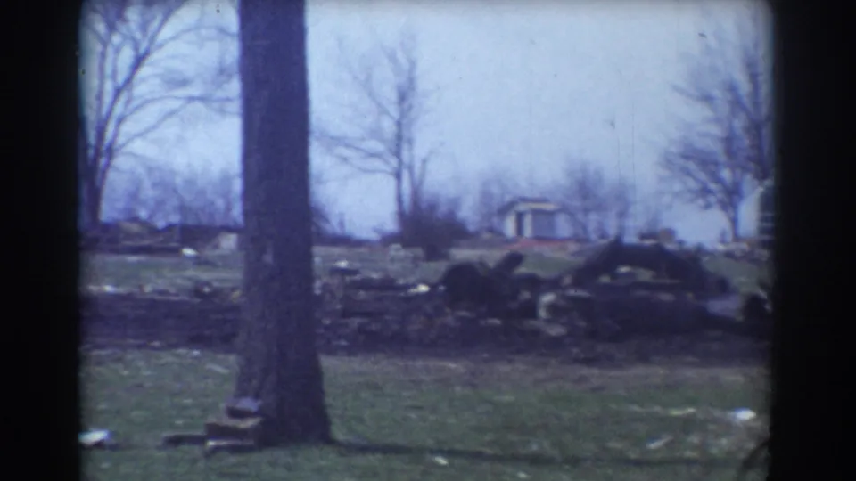 1958 York, Nebraska 8mm frame showing a bare tree, scattered yard debris, logs, and distant trailers across a grassy field.