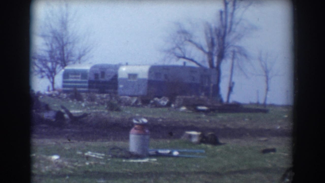 Trailers and a mobile home beyond a grassy yard with a milk can and bare trees.