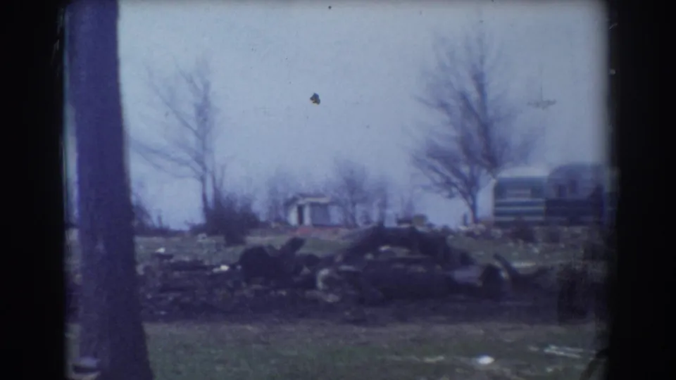1958 York, Nebraska frame with a foreground tree trunk, logs, a small shed, bare trees, and distant trailers in a grassy yard.