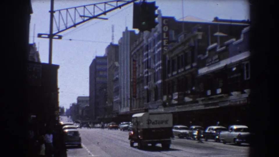 1967 Brisbane downtown street scene with classic cars, pedestrians, overhead signals, shopfront signage, and a delivery truck.