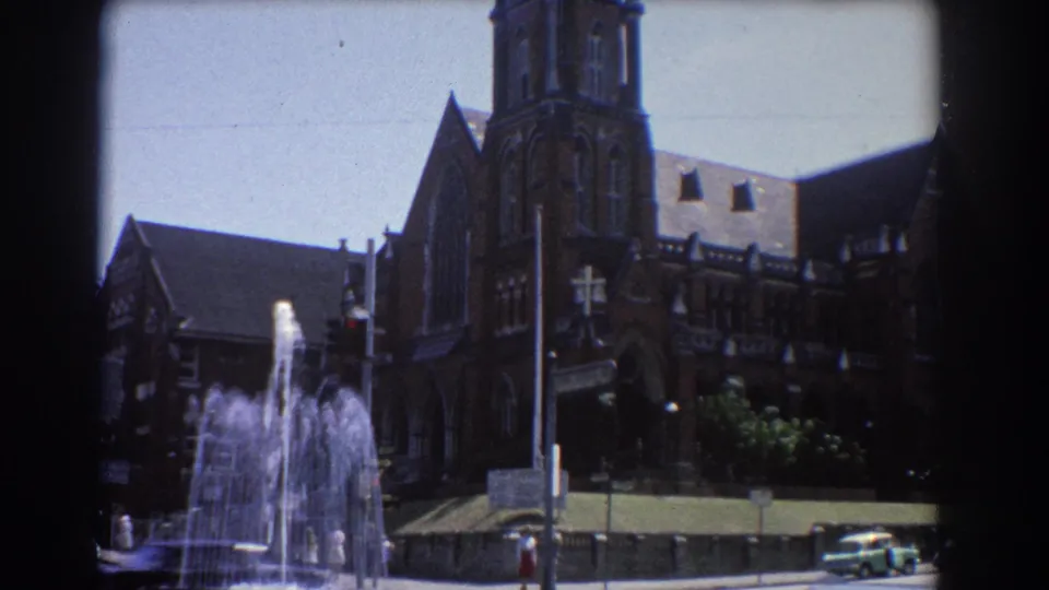 1967 Brisbane street view with a water fountain, brown brick cathedral-like building, traffic pole, parked car, and bright sky.