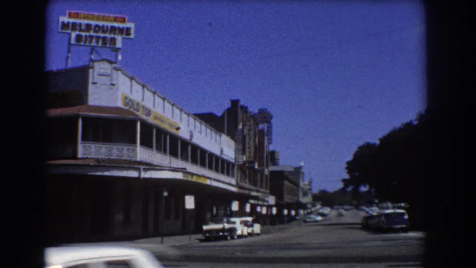 1967 Brisbane street view with a Melbourne Bitter sign above commercial buildings, parked mid-century cars, road, and clear blue sky.