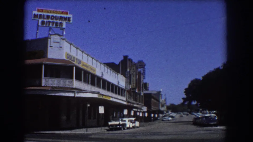 Brisbane 1967 home-movie frame looking down a commercial street with rooftop signage, awnings, parked cars, and deep blue sky.