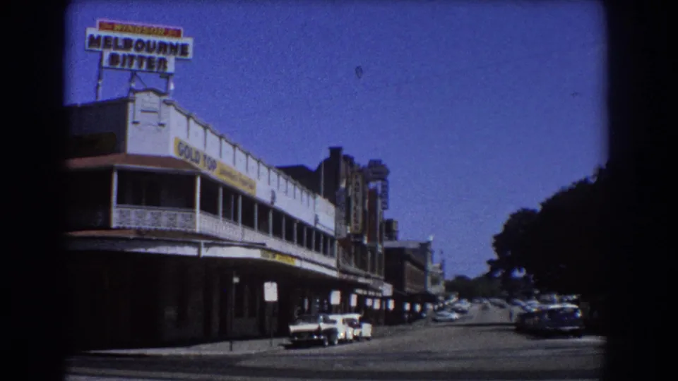 1967 Brisbane street view showing a corner commercial building with Melbourne Bitter and Gold Top signage and vintage cars under a clear sky.