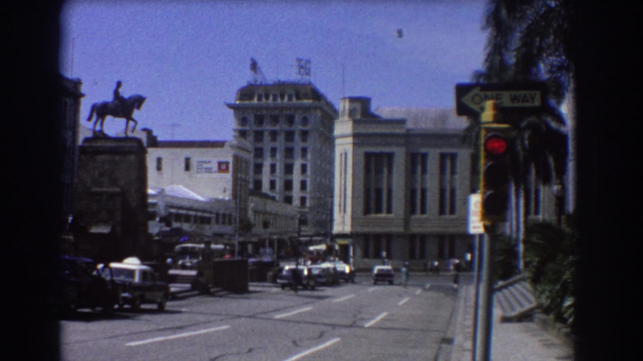 City intersection with cars, a red traffic light, a one-way sign, palm trees, and a large building with a T&G sign.