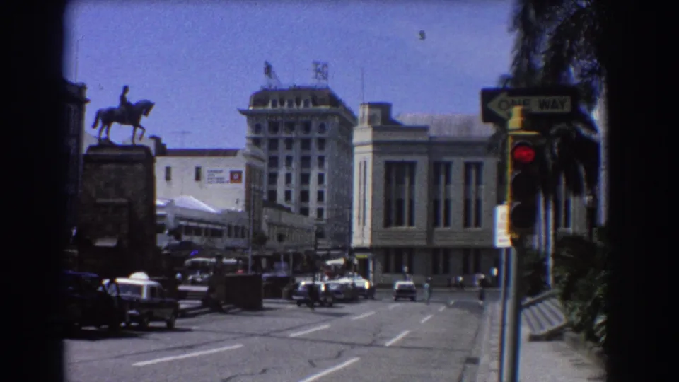 1967 Brisbane city-center traffic scene with cars crossing an intersection, a red light, one-way sign, palms, and a visible T&G sign.